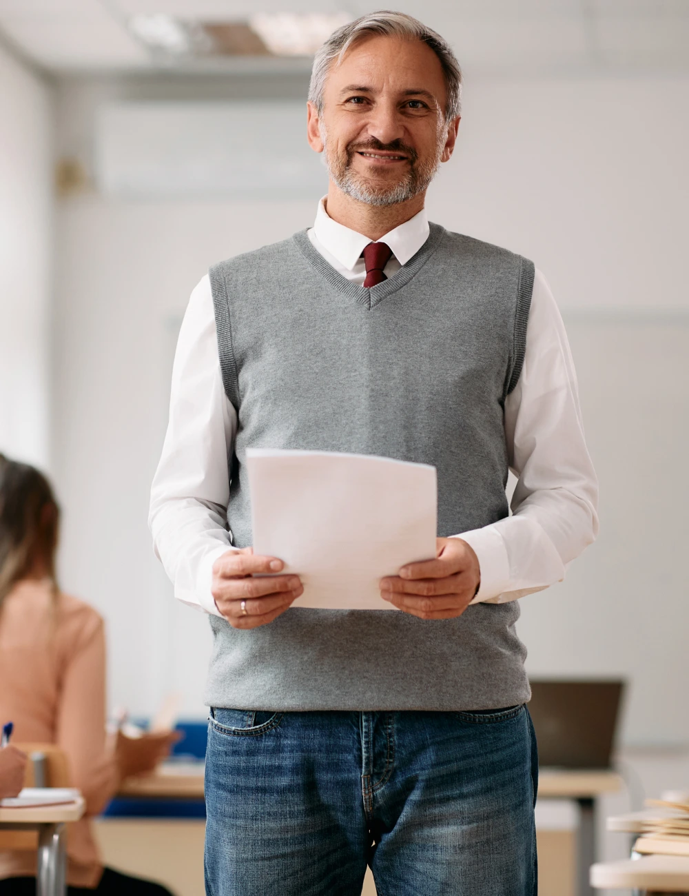 Smiling middle-aged man holding papers in a classroom setting, wearing a gray sweater vest, white shirt, and red tie.