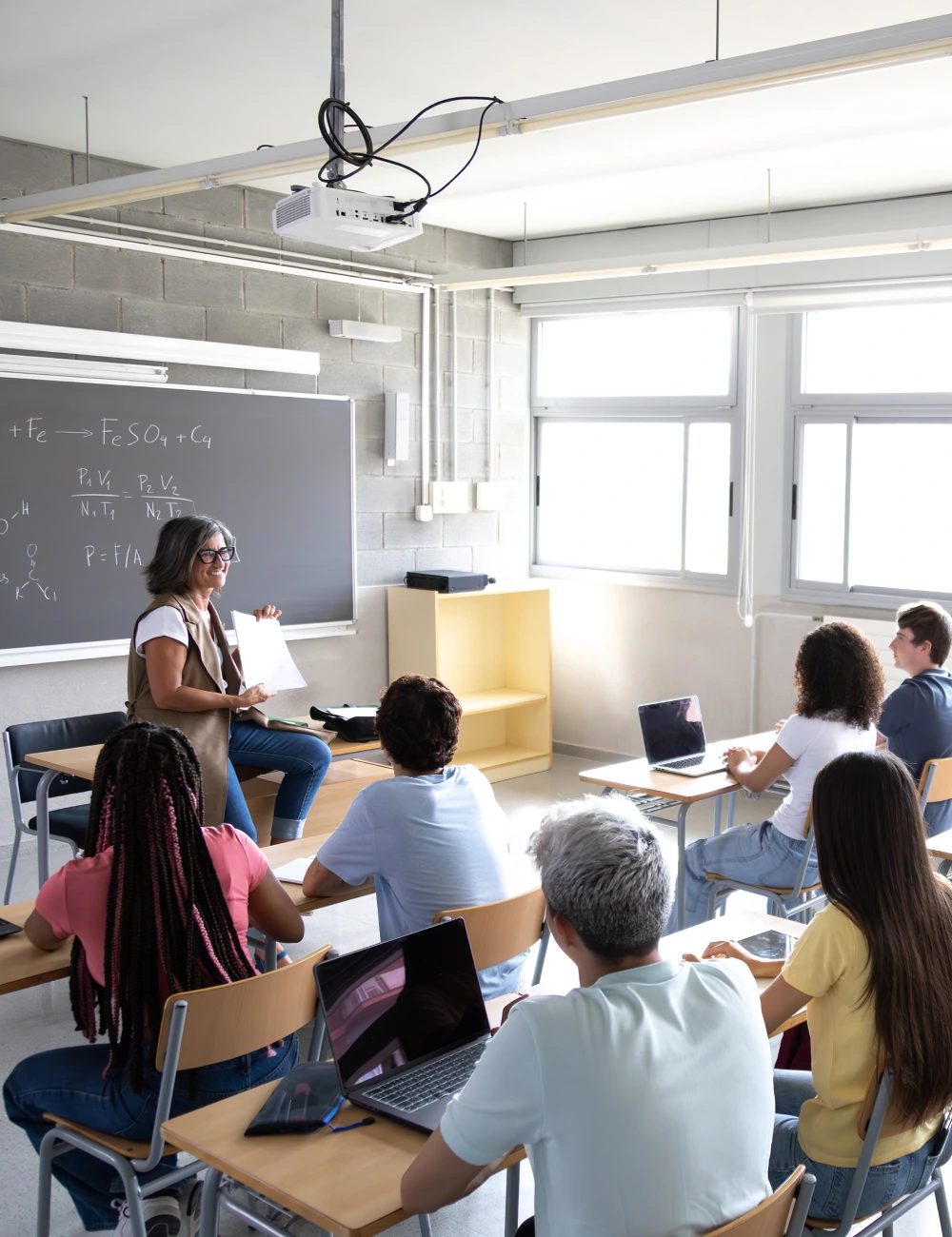 Teacher sitting on desk holding paper and smiling at diverse students seated in classroom with laptops and tablets.