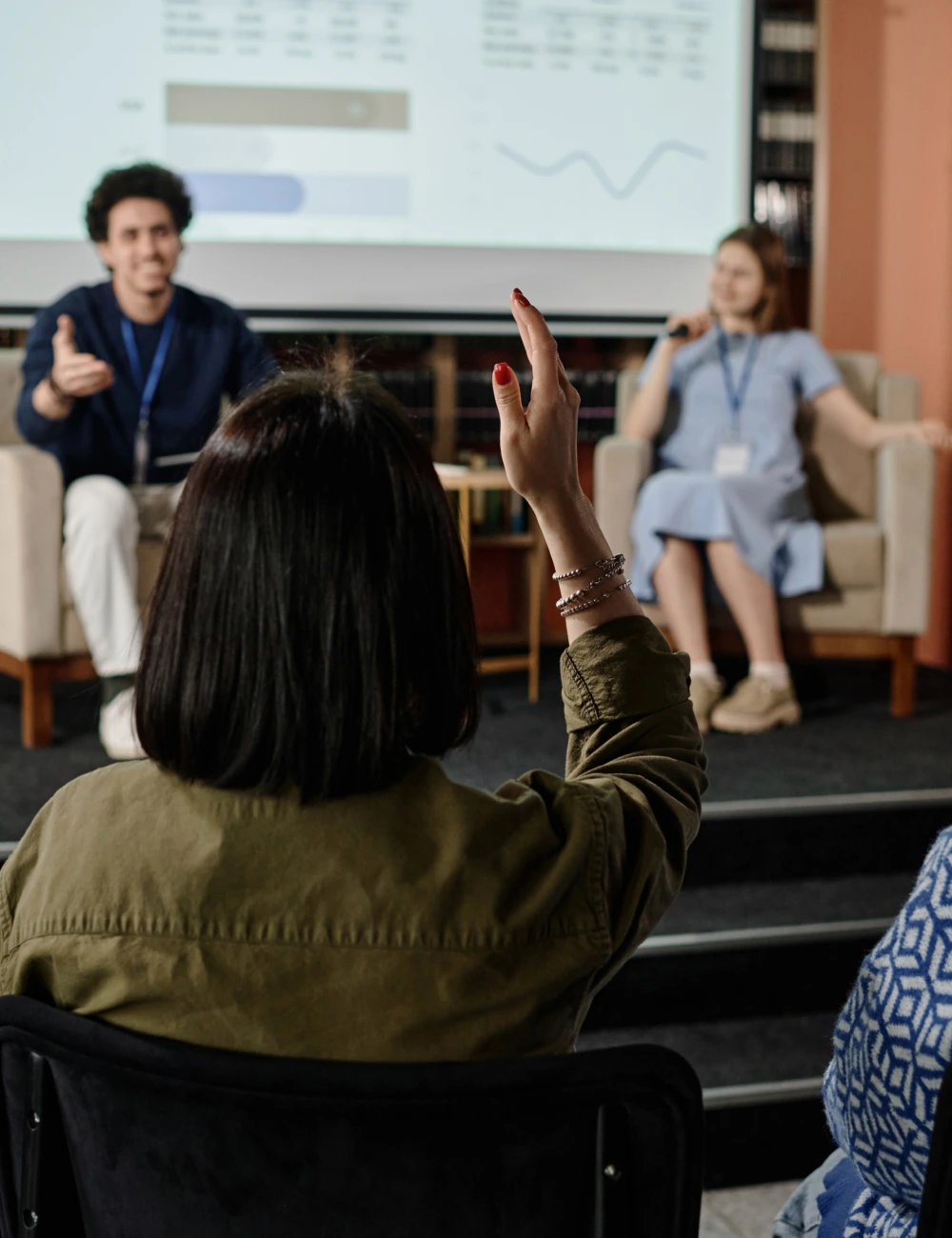 Person raising hand in an audience while two speakers sit on a stage with a presentation screen behind them.