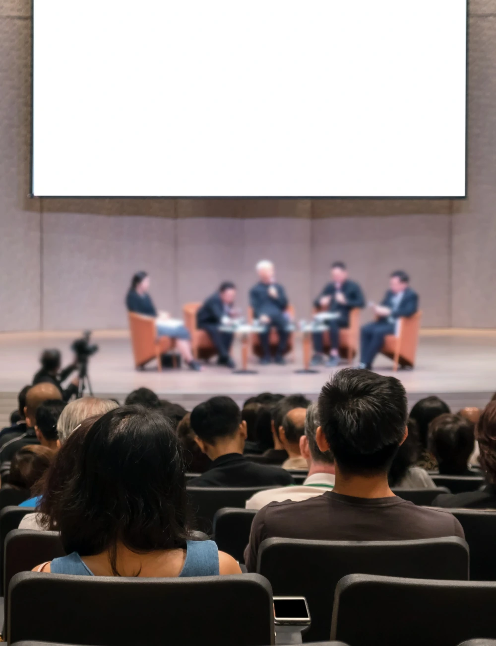 Audience seated facing a panel of five speakers on stage with a large blank screen behind them in a conference or seminar setting.