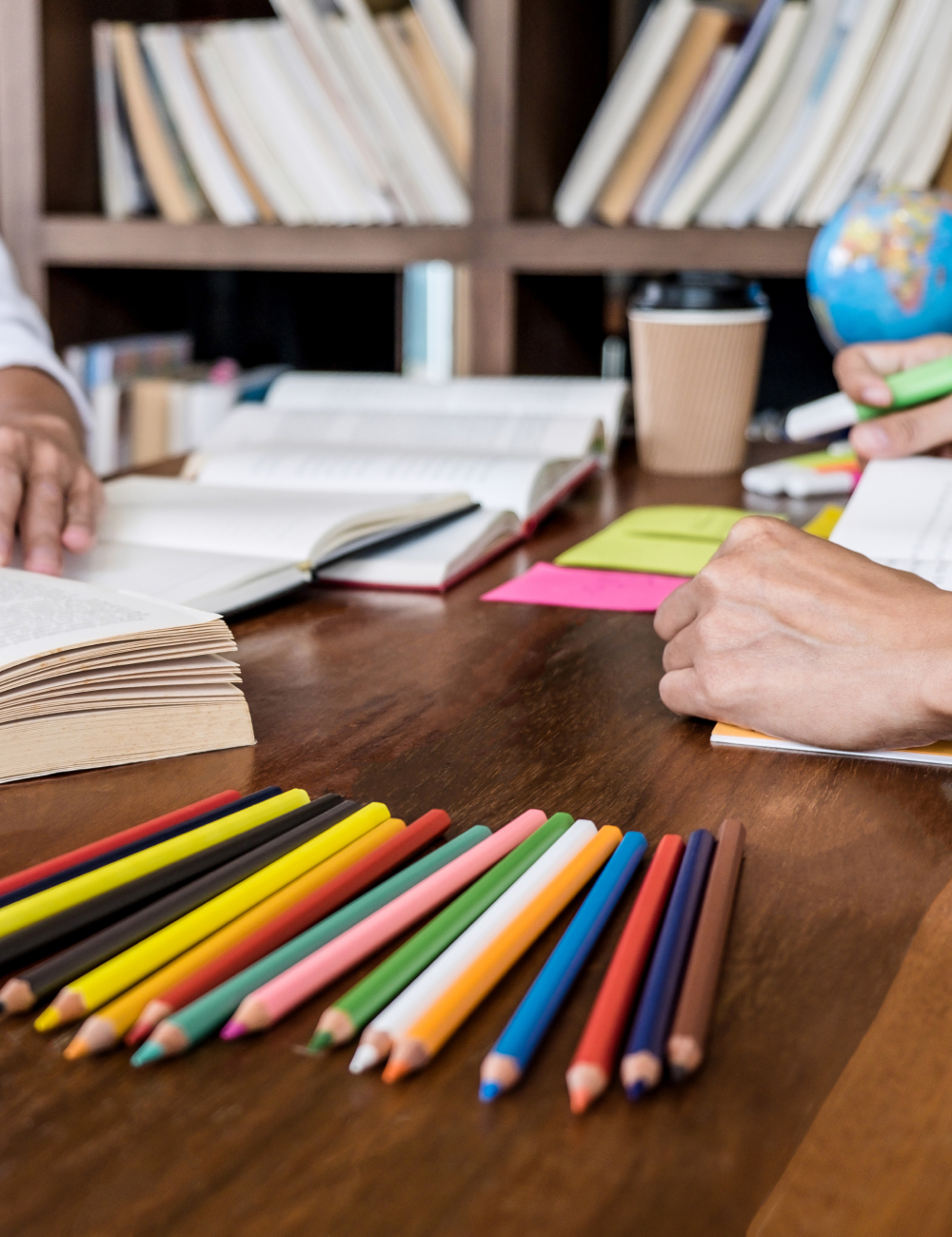 Colorful pencils lined up on a wooden table with open books, sticky notes, and a person writing with a green highlighter in a study setting.