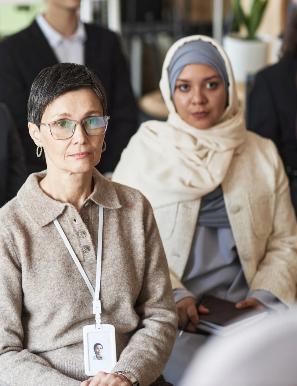 Two women seated in a professional setting, one wearing glasses and a name badge, the other wearing a light hijab and holding a notebook and pen.