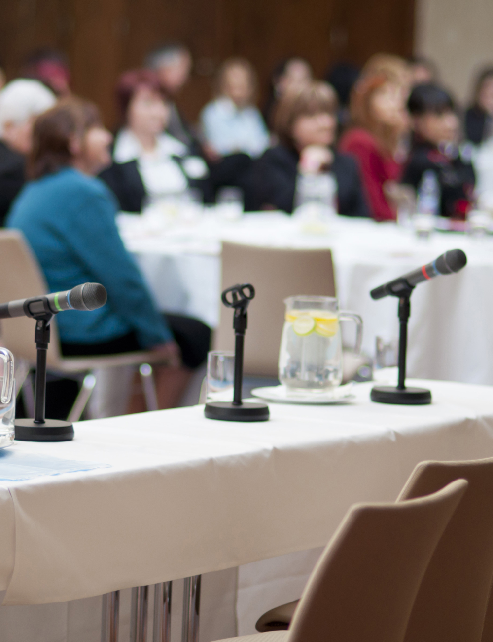 Conference table with microphones and a pitcher of water with lemon slices, blurred audience in background.