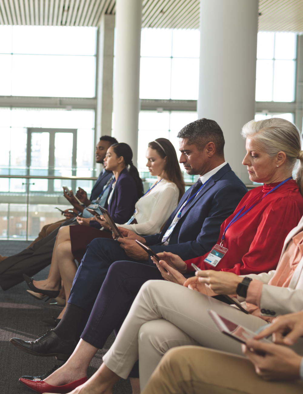 Diverse group of professionally dressed people sitting in a row indoors, all focused on their smartphones or tablets.