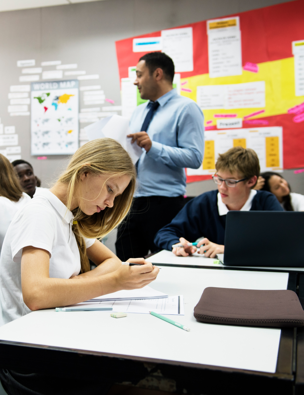Students focused on writing and working at desks while a teacher stands near a colorful classroom bulletin board.
