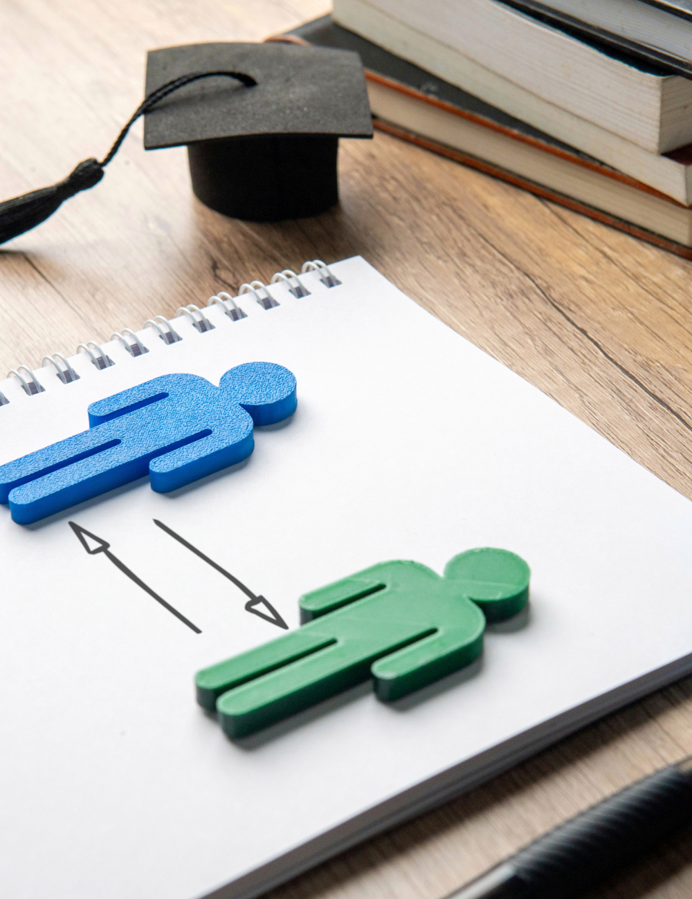 Blue and green human figures on a notebook with double-headed arrows between them, next to a graduation cap and stacked books on a wooden desk.