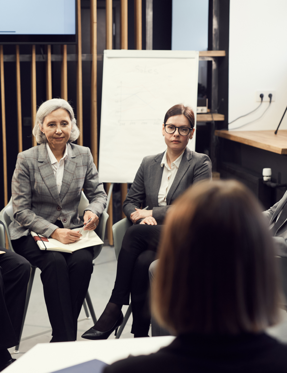 Two businesswomen in gray suits attentively listening during a meeting with a flip chart in the background.