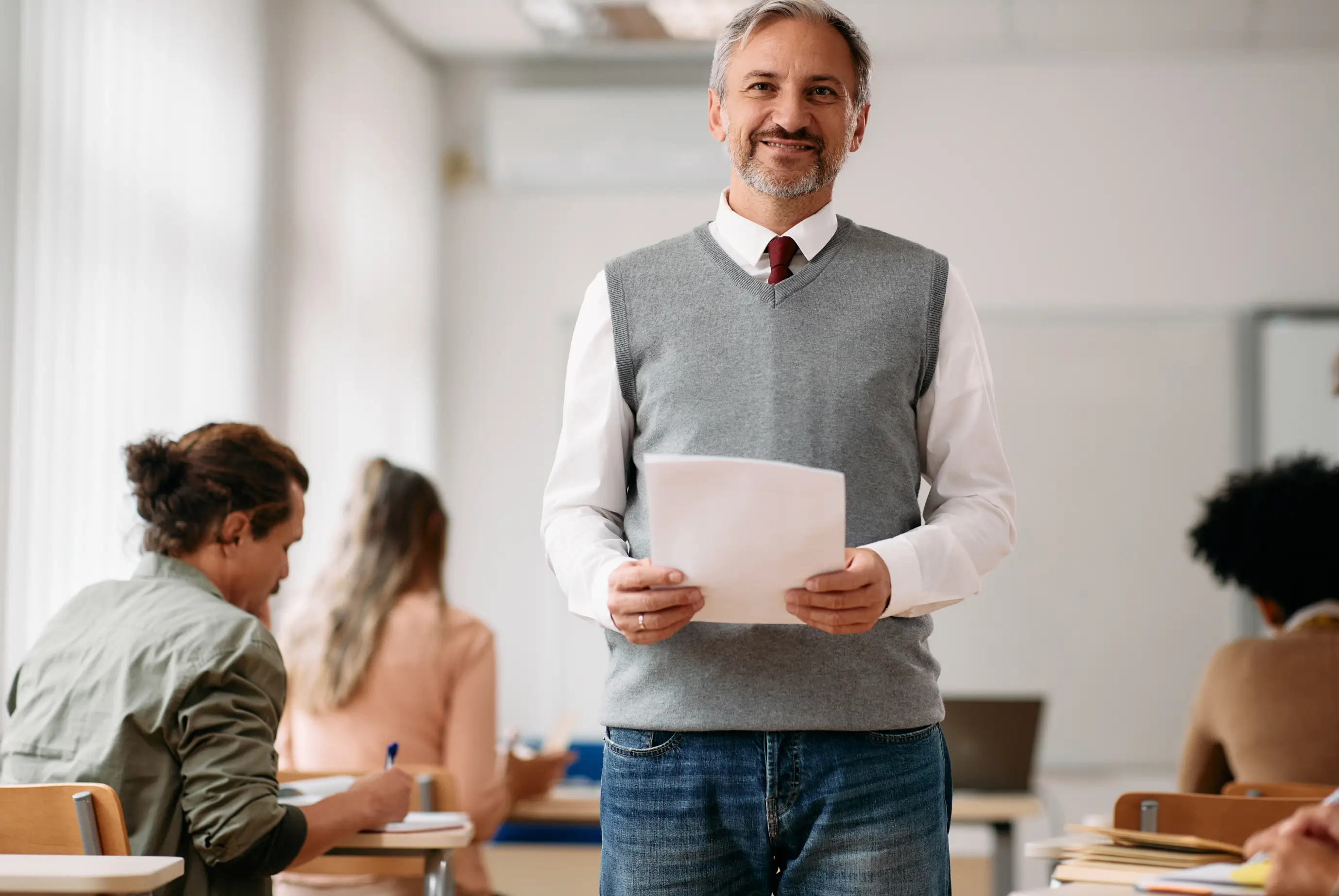 Smiling teacher in gray vest and white shirt holding papers in a classroom with students seated at desks.