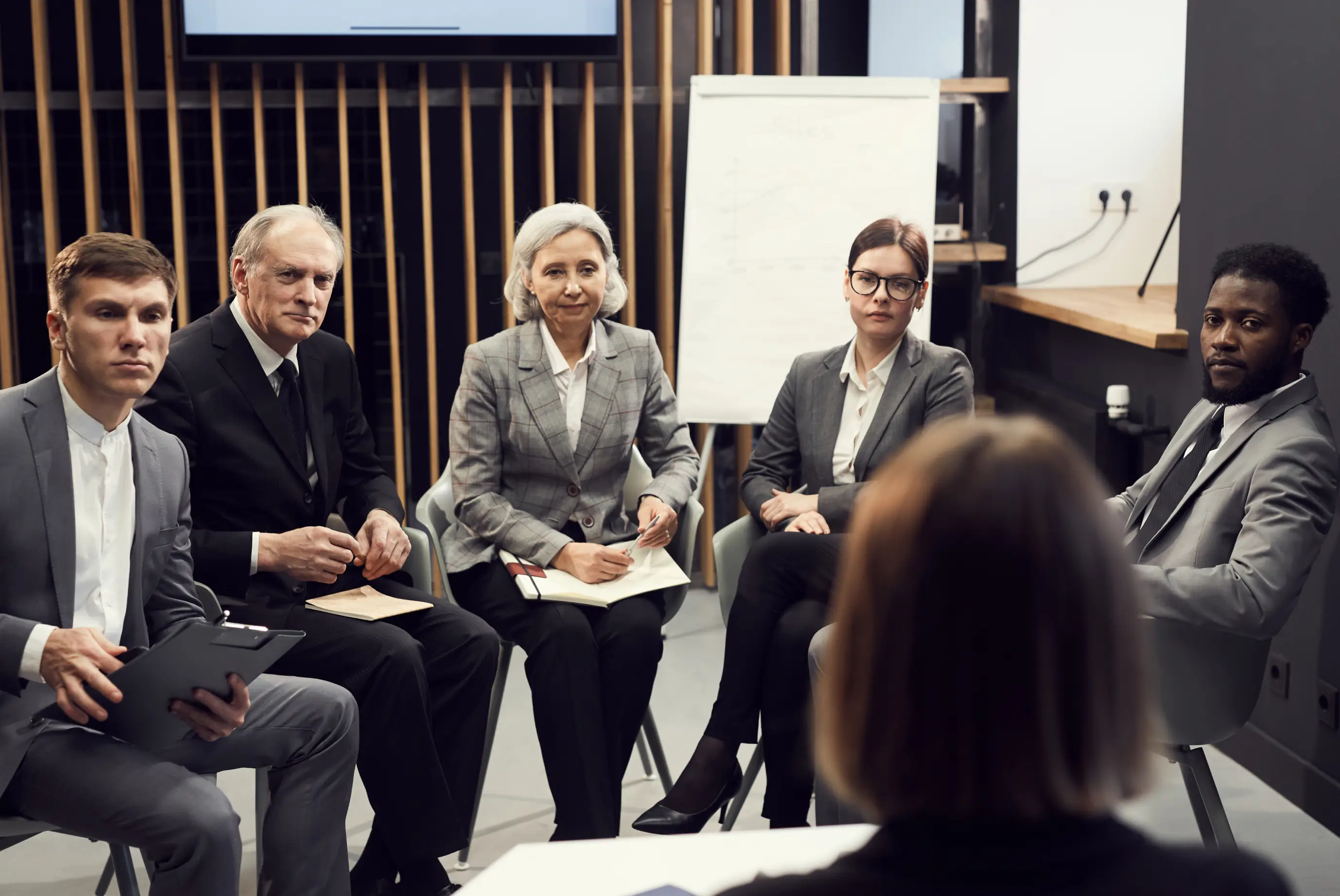 Five professionals in business attire sitting in a meeting room, attentively listening to a person with blurred back facing them.