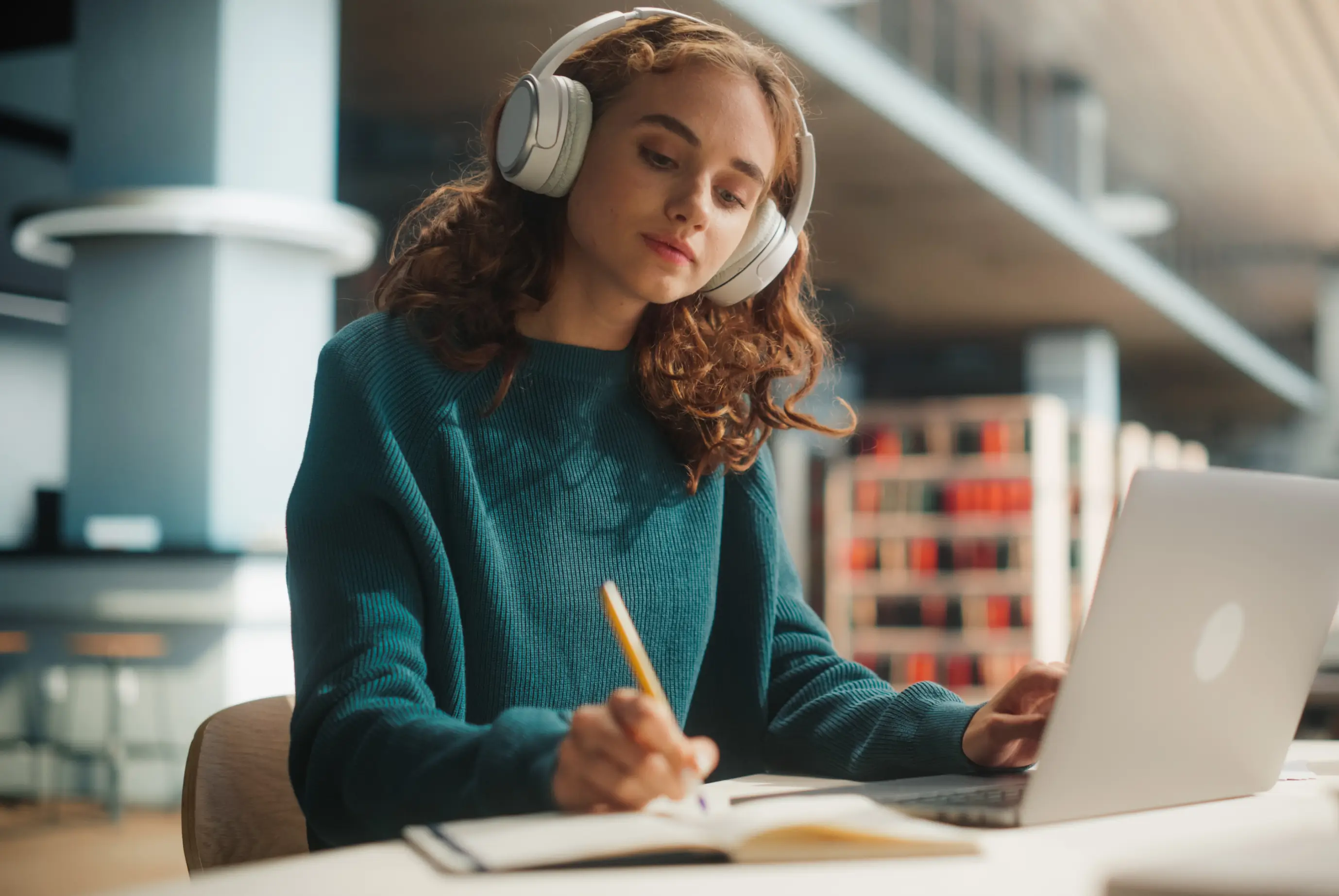 Young woman wearing headphones writing in a notebook while using a laptop in a library.