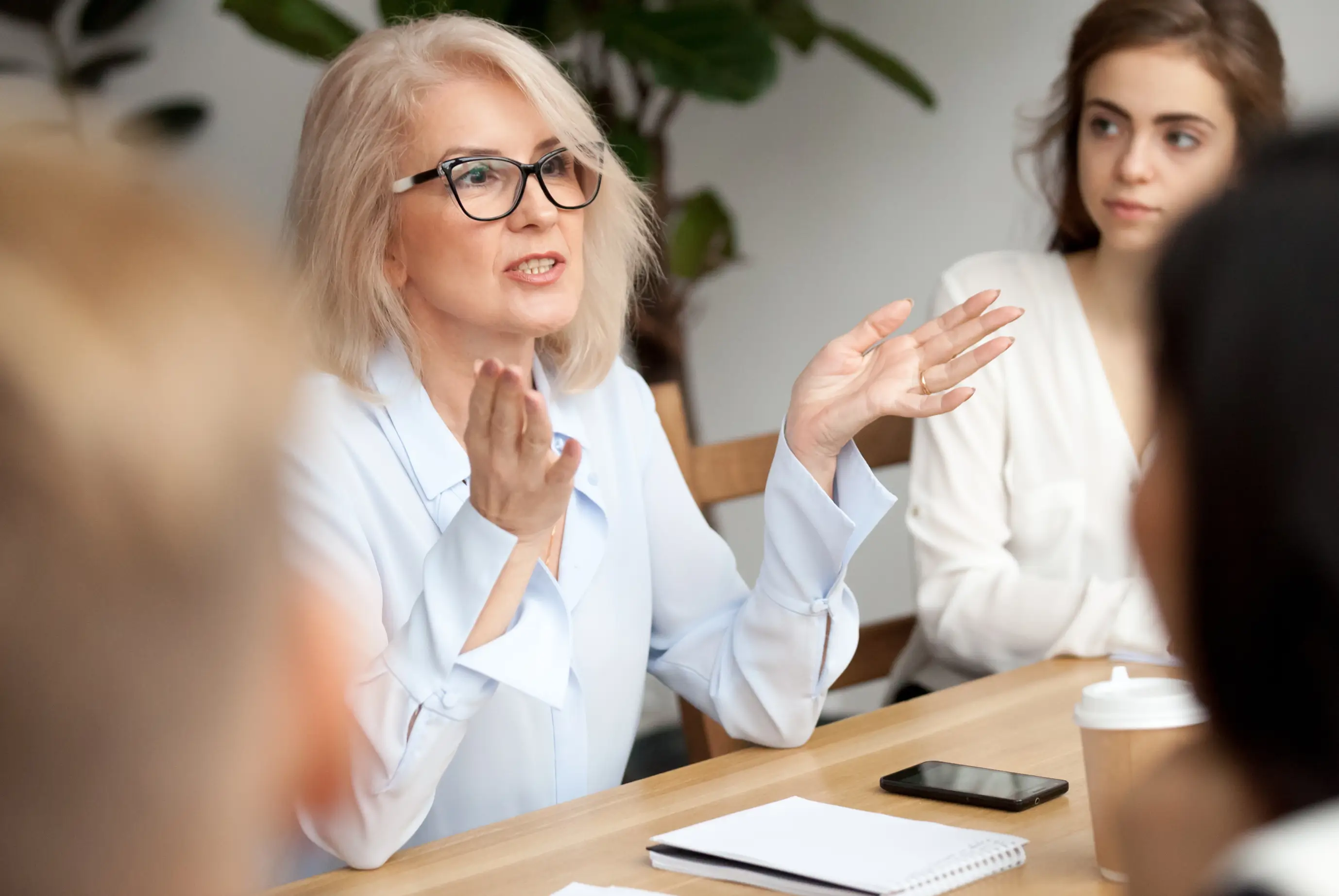 Older woman with glasses speaking and gesturing during a meeting at a table with notebooks and coffee.